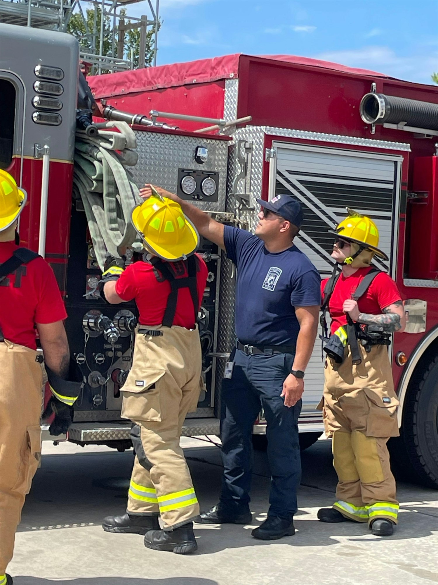Firefighters in training are interacting near a fire truck, preparing equipment and discussing procedures.