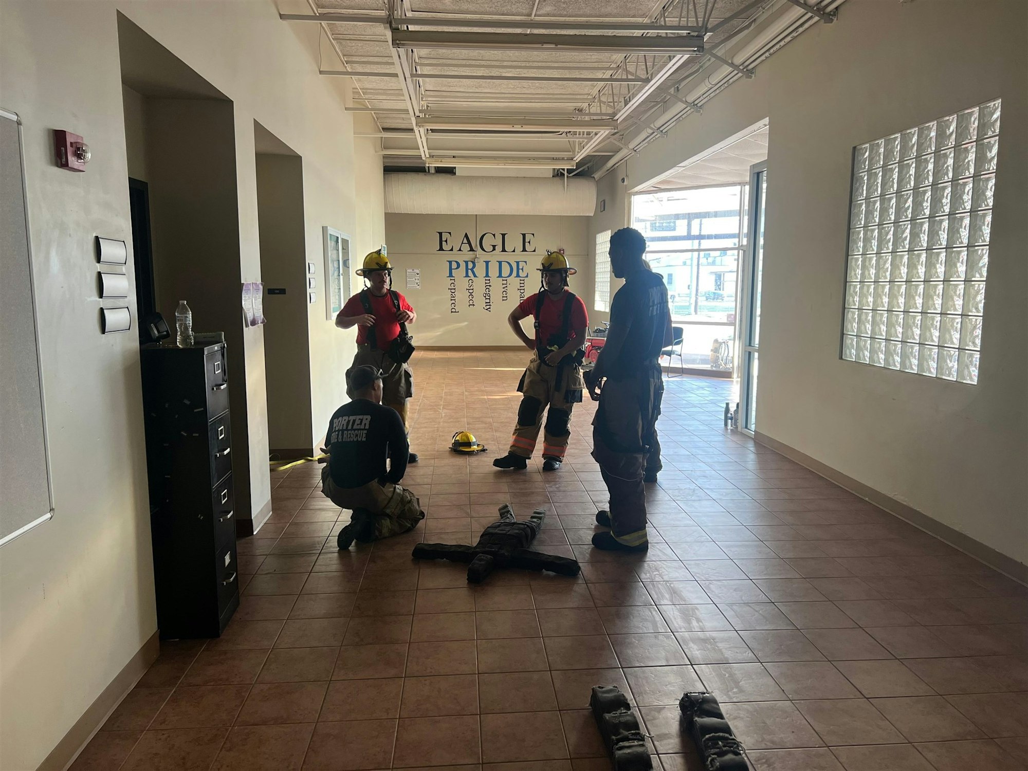 Firefighters in gear are gathered indoors, discussing or preparing for a training or emergency response. "Eagle Pride" is on the wall.