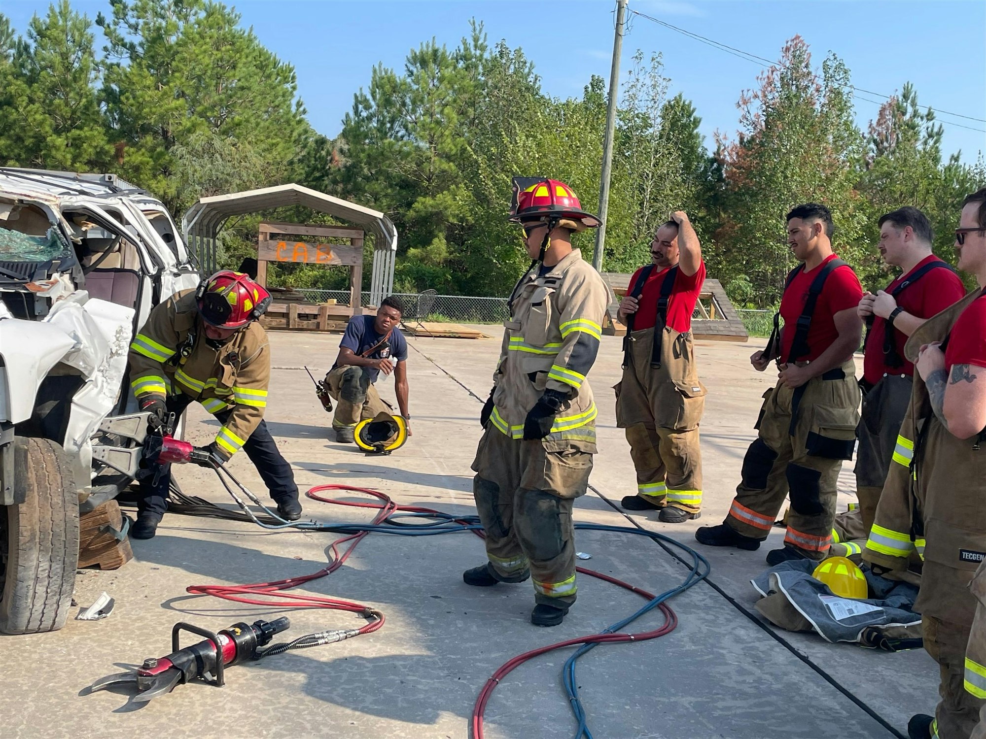 Firefighters conducting a training exercise using hydraulic tools on a vehicle, with onlookers observing the process.