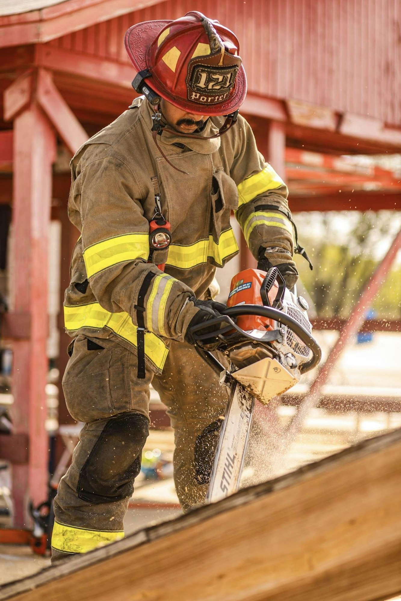 A firefighter in protective gear uses a chainsaw to cut wood, with sawdust flying in the air.
