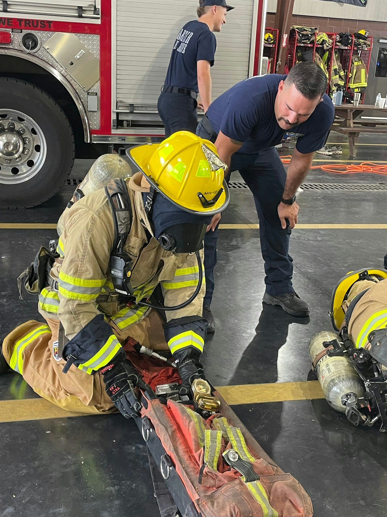 A firefighter practices equipment handling, while fellow firefighters look on in a training facility.