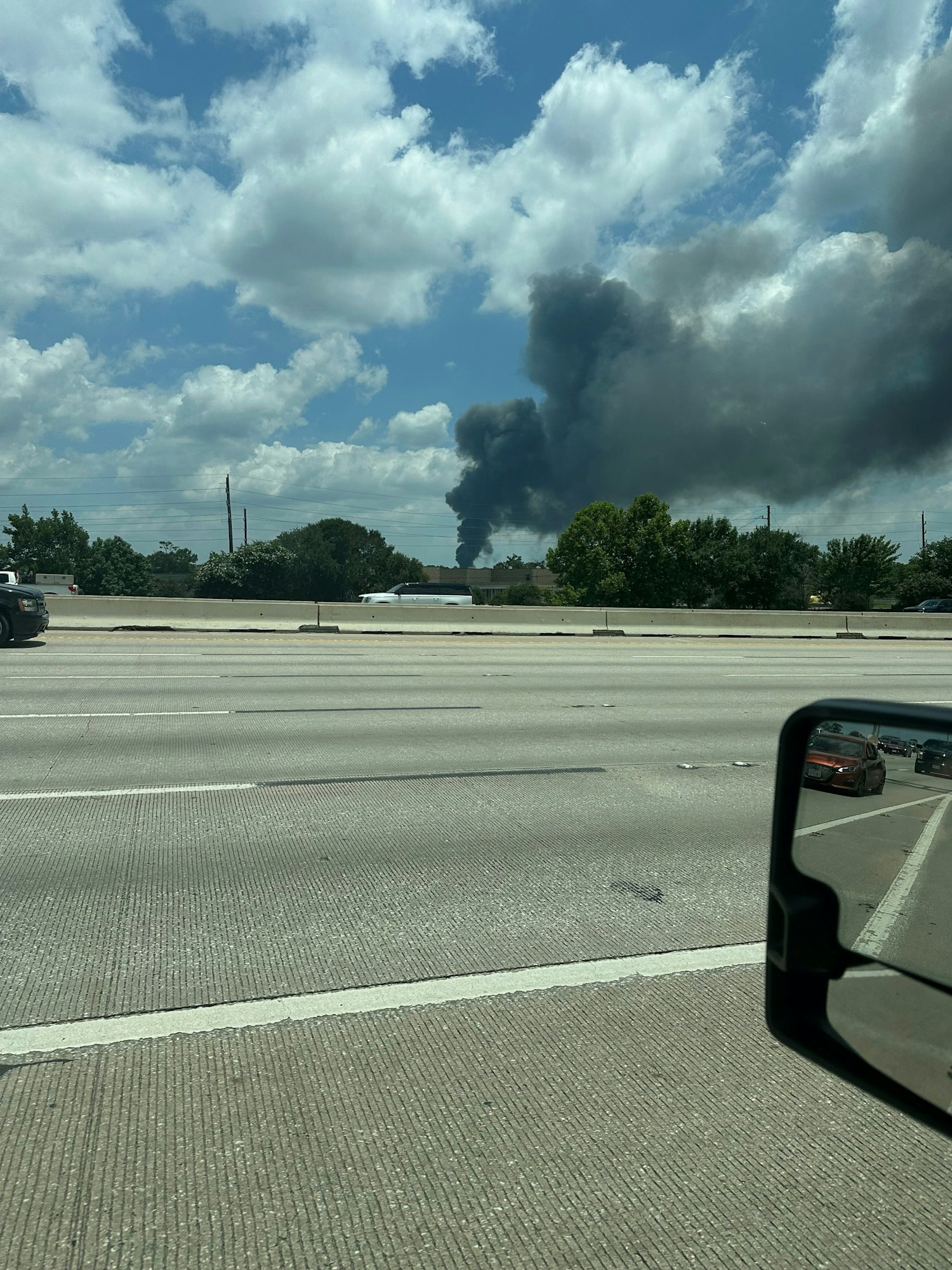 Large plume of dark smoke visible over highway, possibly from a distant fire, under a partly cloudy sky.
