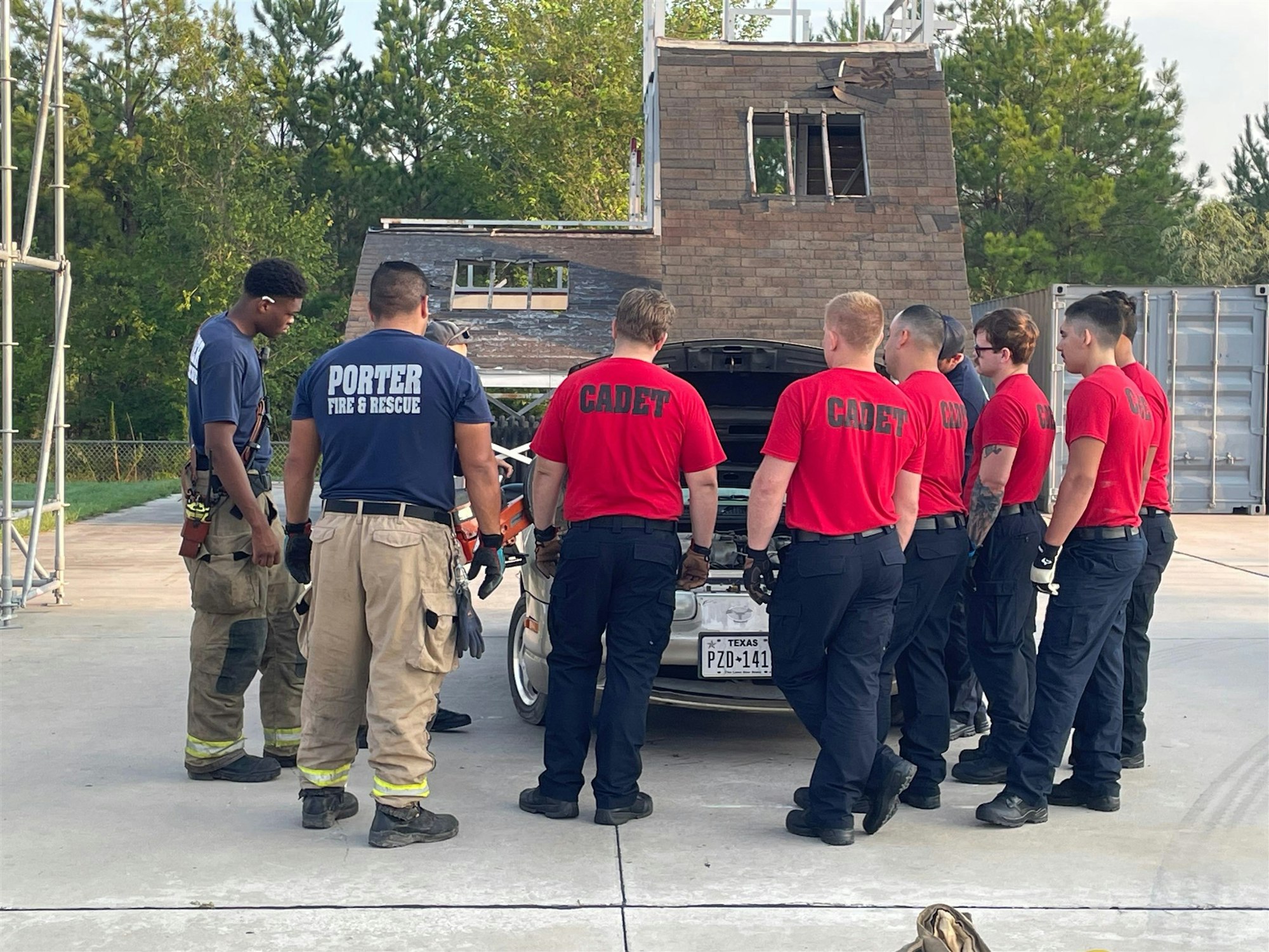 Firefighters and cadets are gathered around a car, possibly training for rescue operations, with a damaged structure in the background.