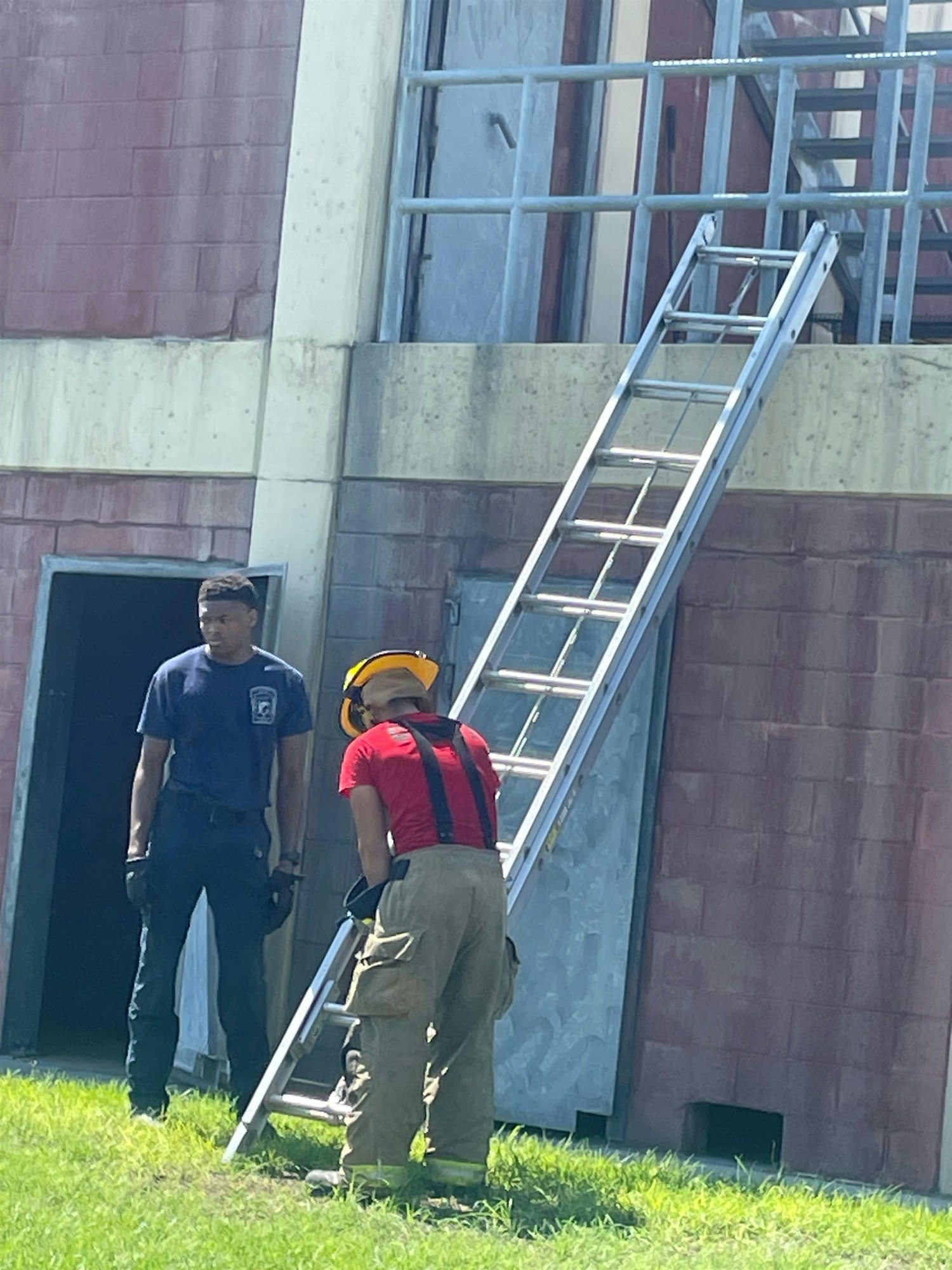 Two individuals are near a ladder; one is in firefighter gear, while the other stands nearby, possibly in training or observation.