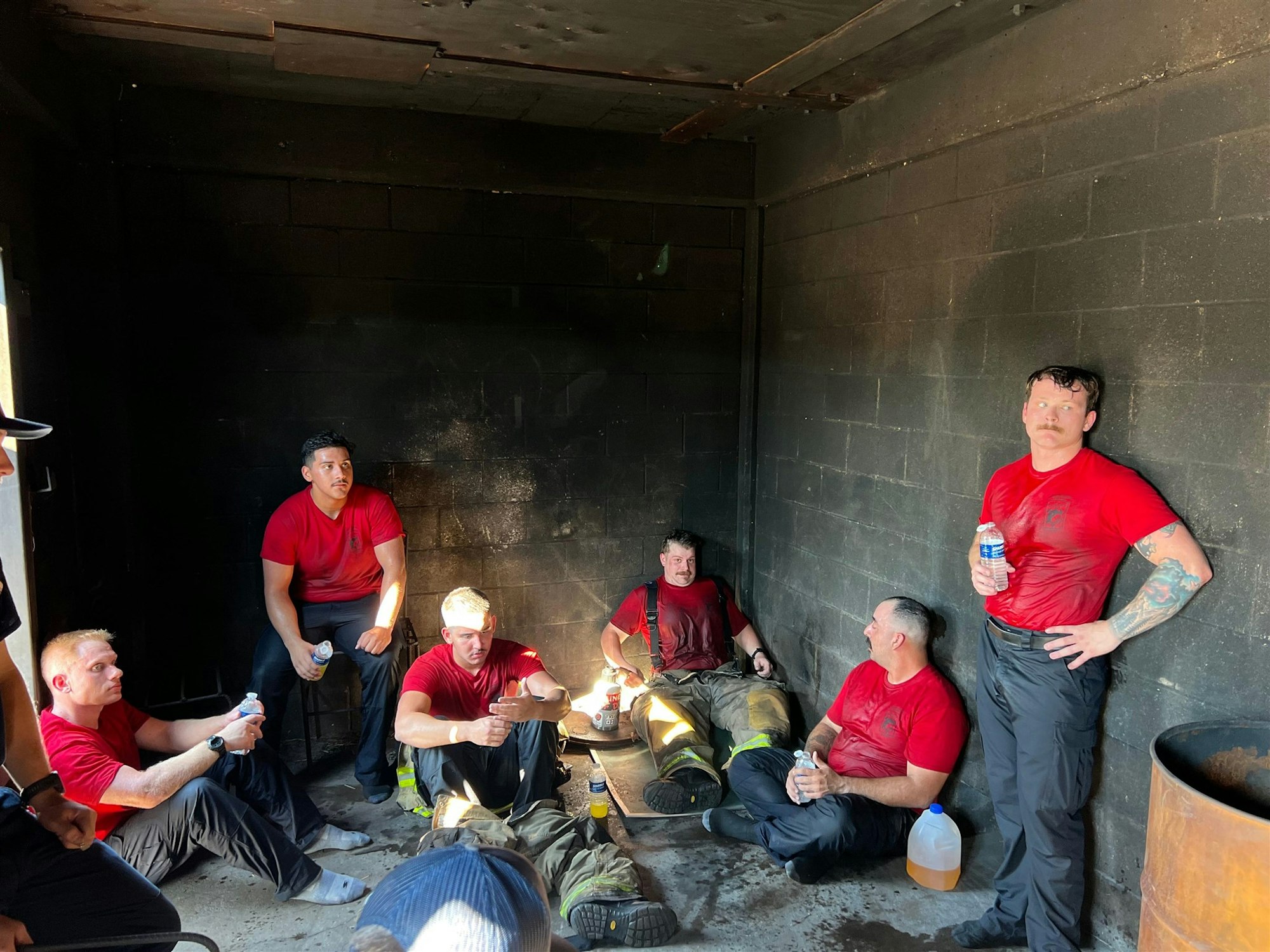 A group of individuals in red shirts rests in a dimly lit room, some drinking water, appearing fatigued after a task or training.