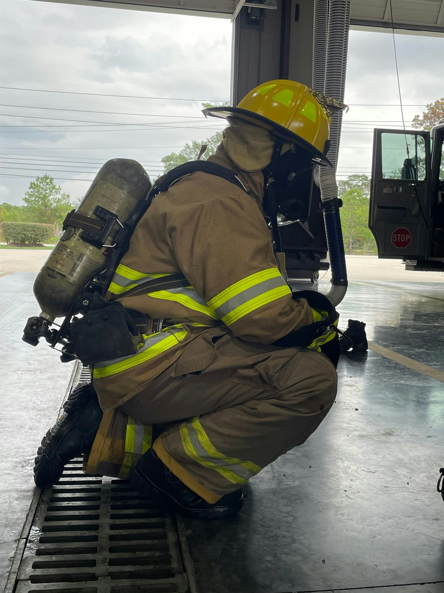 A firefighter in gear, including a helmet and air tank, kneels in a fire station with a truck visible in the background.