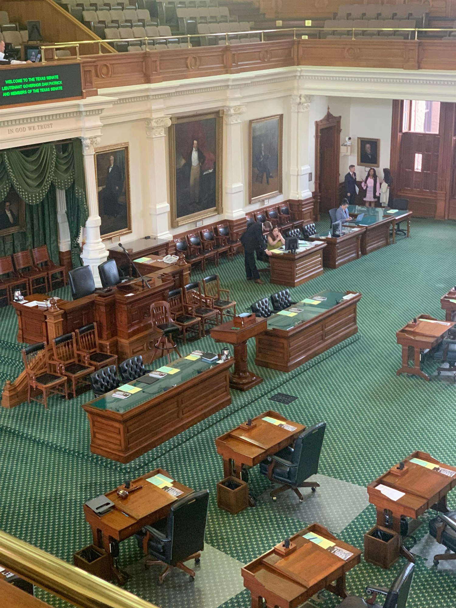 An overview of a legislative chamber with wooden desks, chairs, and portraits on the walls, likely during a session.