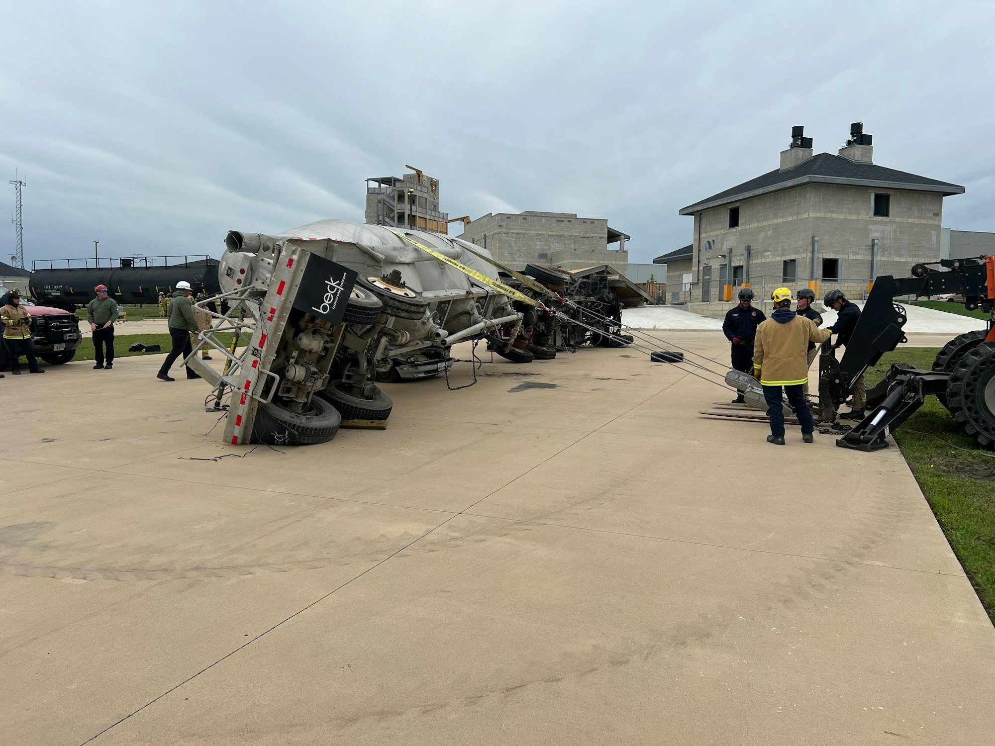 An overturned semi-trailer, emergency responders on site, with industrial and residential buildings in the background.