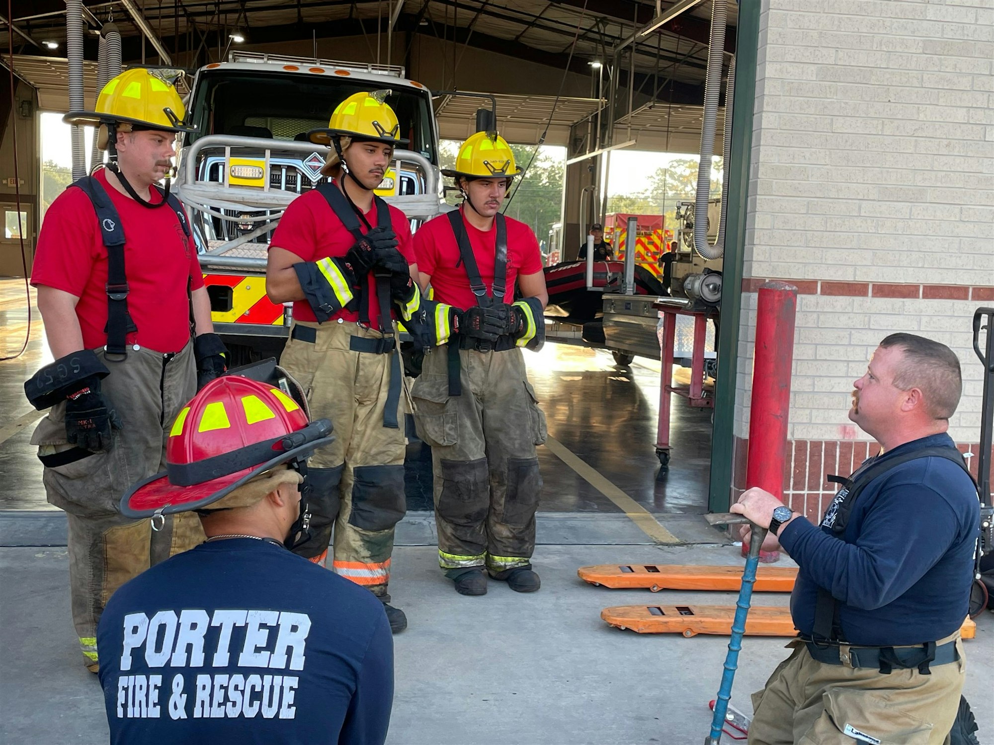 Firefighters in training engaged in discussion at a fire station, with a fire truck visible in the background.