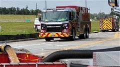 The image shows a fire truck on a road, with another truck in the background and equipment visible. Green fields are nearby.