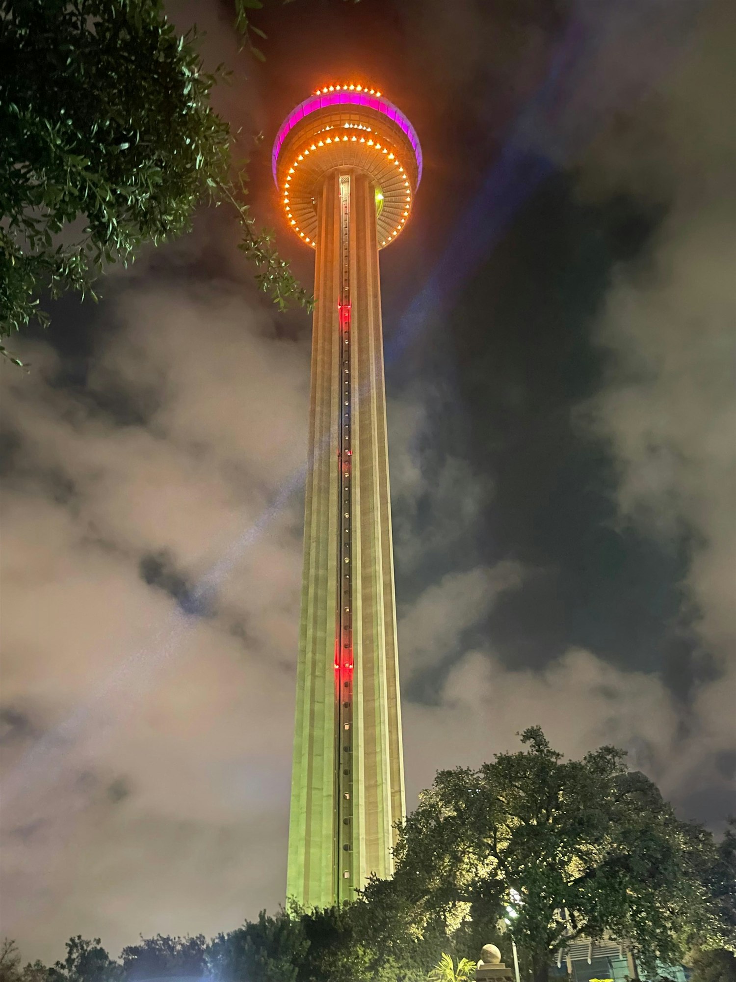 A tall illuminated tower at night, with colorful lights, partially obscured by clouds and surrounded by trees.