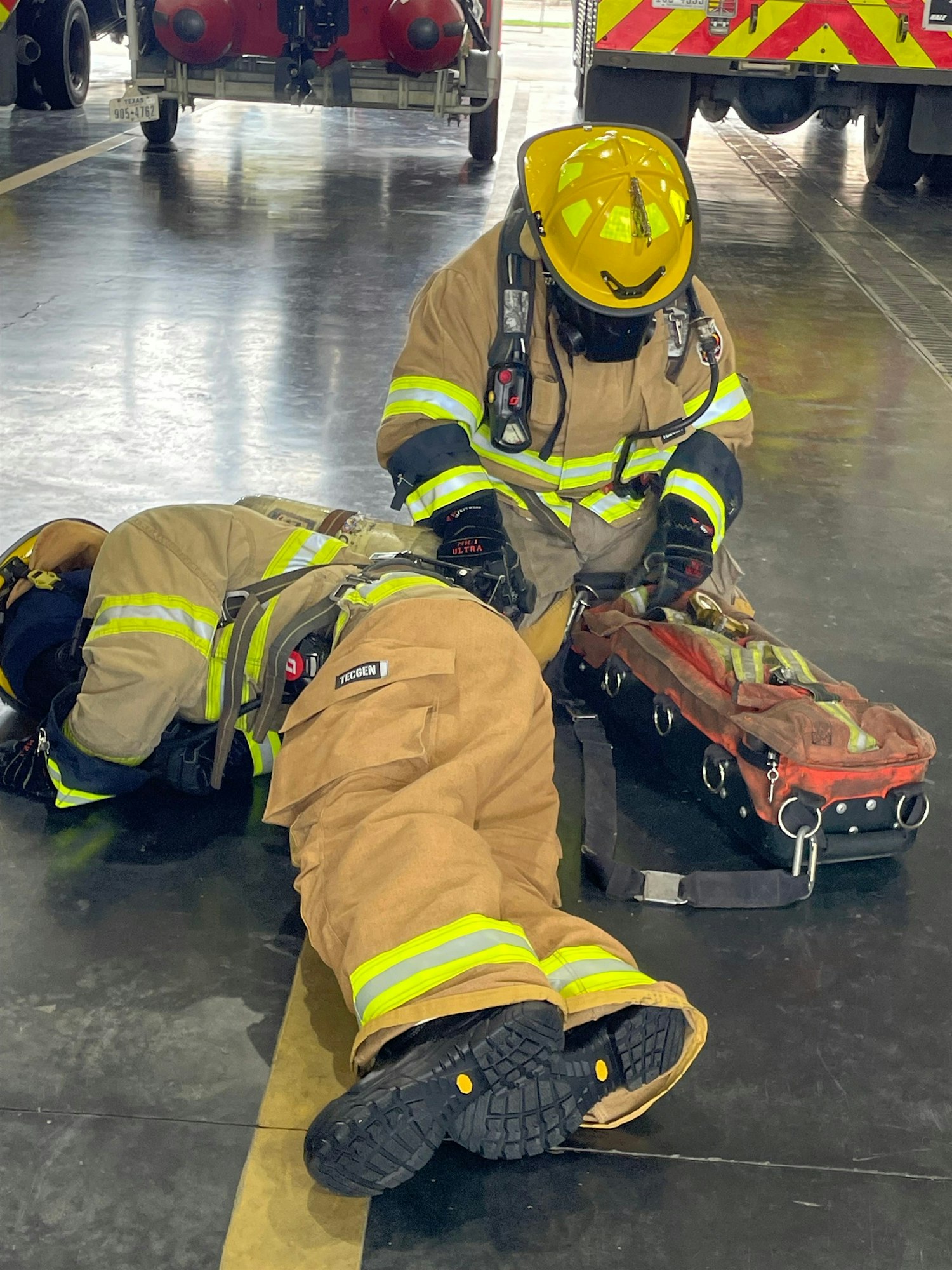 Two firefighters in uniform are practicing a rescue drill on a stretcher in a fire station.