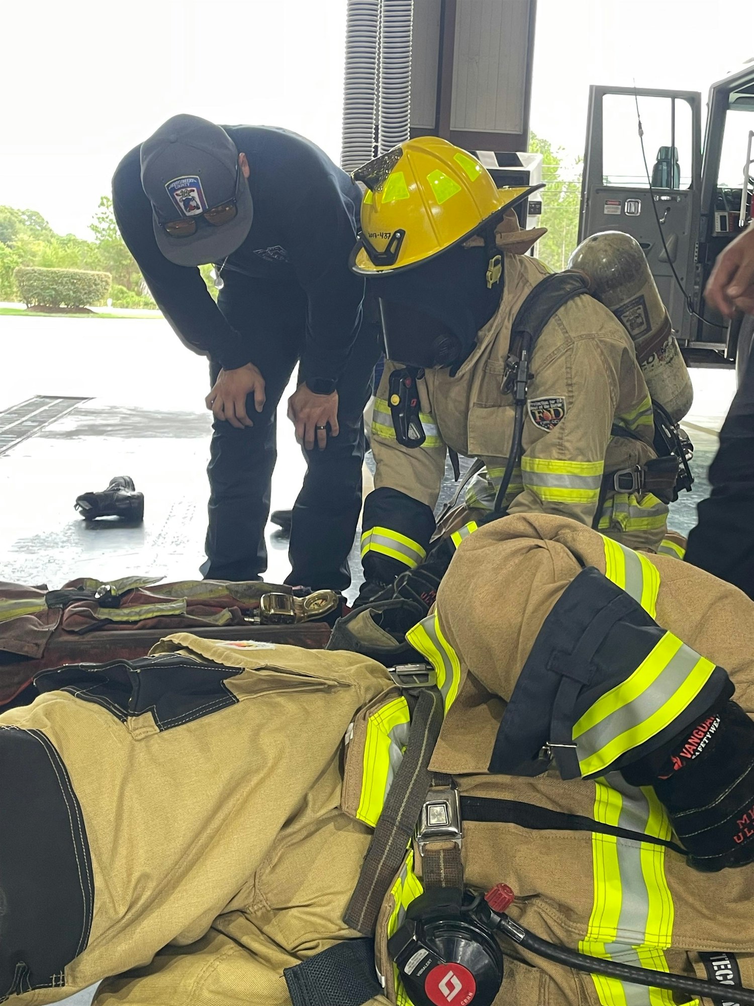 A firefighter in gear is adjusting equipment, while a colleague observes in a fire station setting.