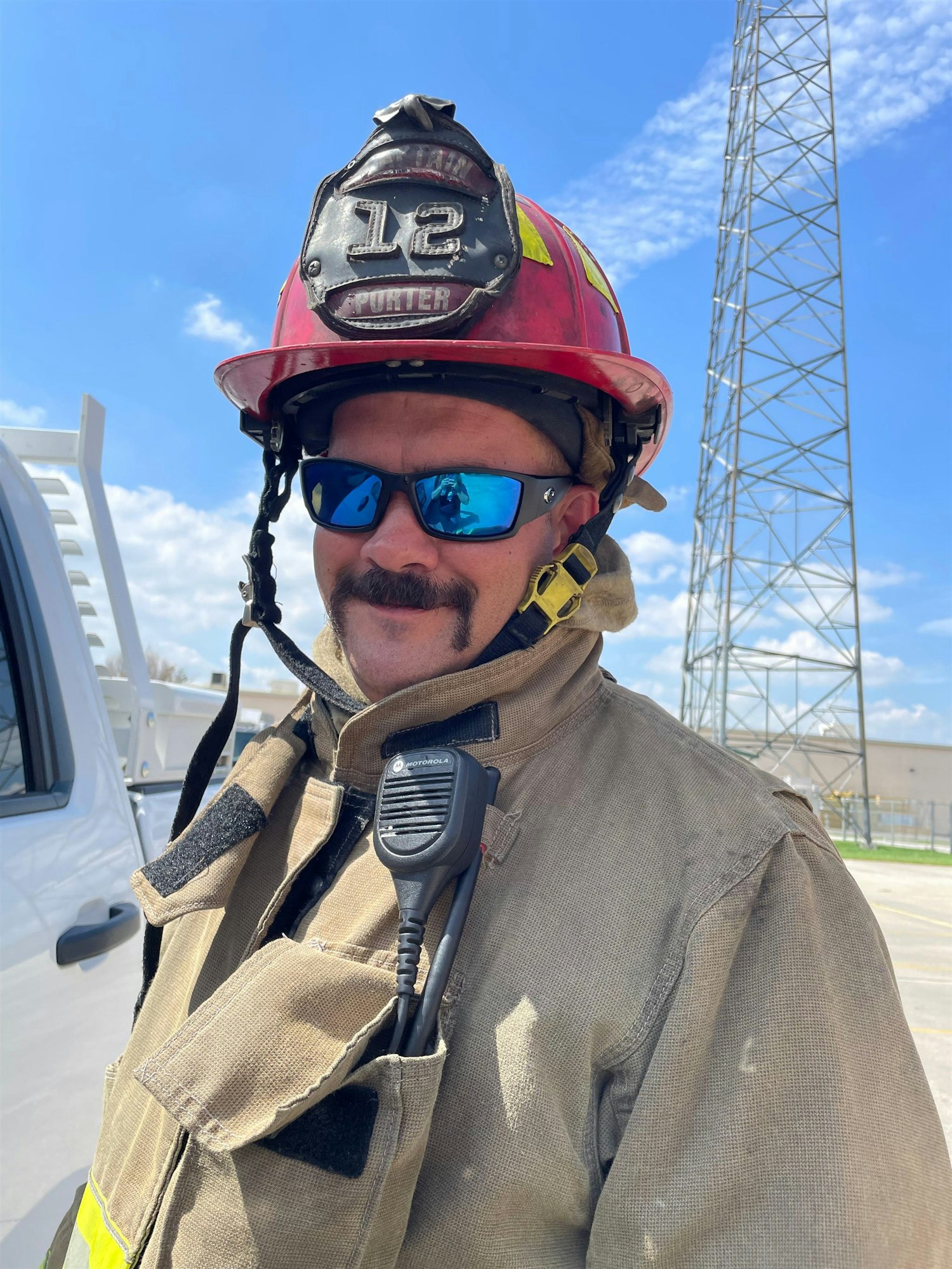 A firefighter in full gear with a helmet, sunglasses, and a radio, posing outdoors under a blue sky.