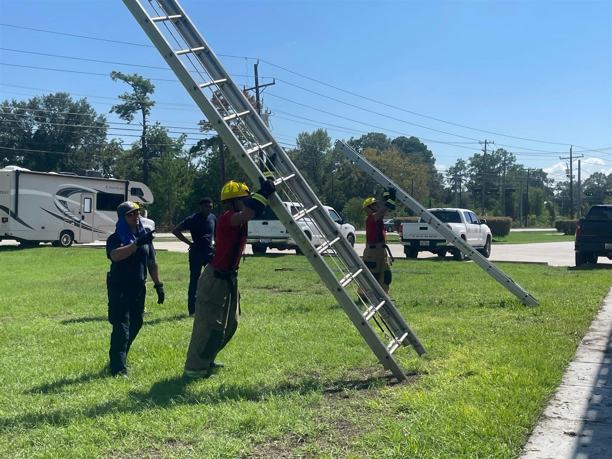 Firefighters are seen working together to carry a ladder on a grassy area near vehicles under a clear blue sky.