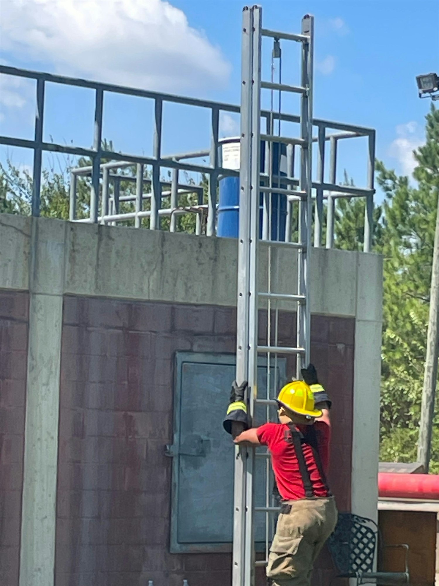 A person in a yellow helmet climbs a ladder against a building, possibly engaged in maintenance or training work.
