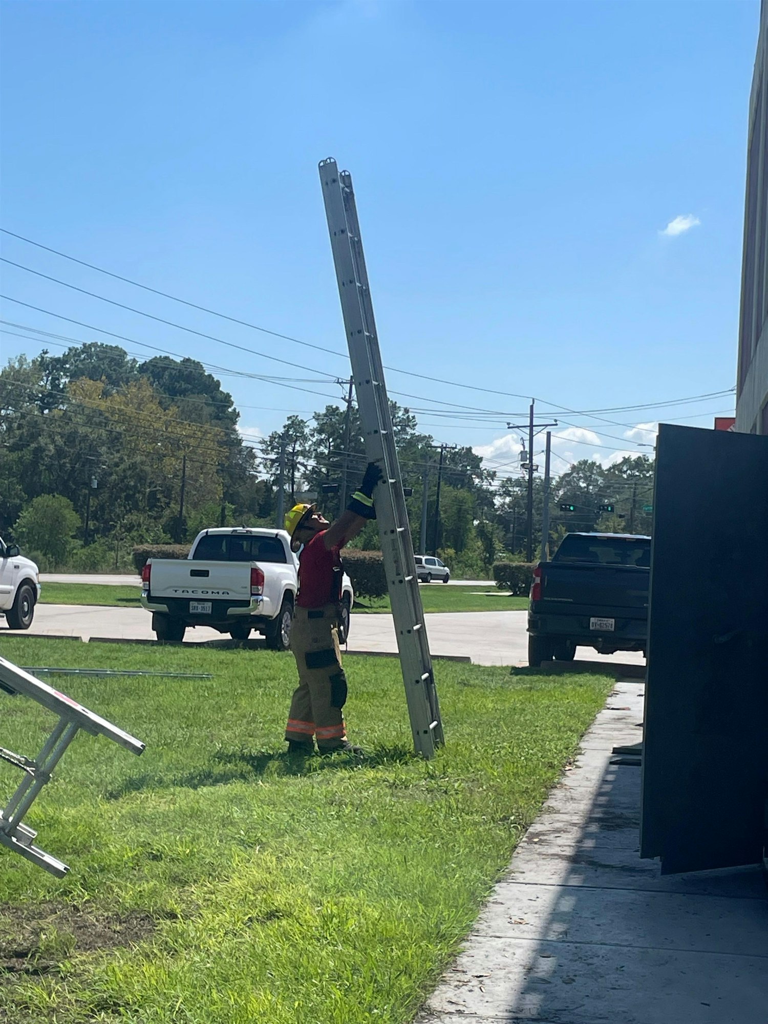 A person in firefighting gear is holding a tall ladder outdoors, with vehicles and trees in the background under a clear sky.