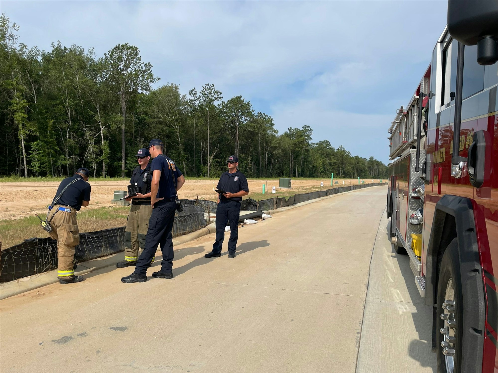 Firefighters are gathered near a fire truck, discussing or inspecting something along a road with a forested background.