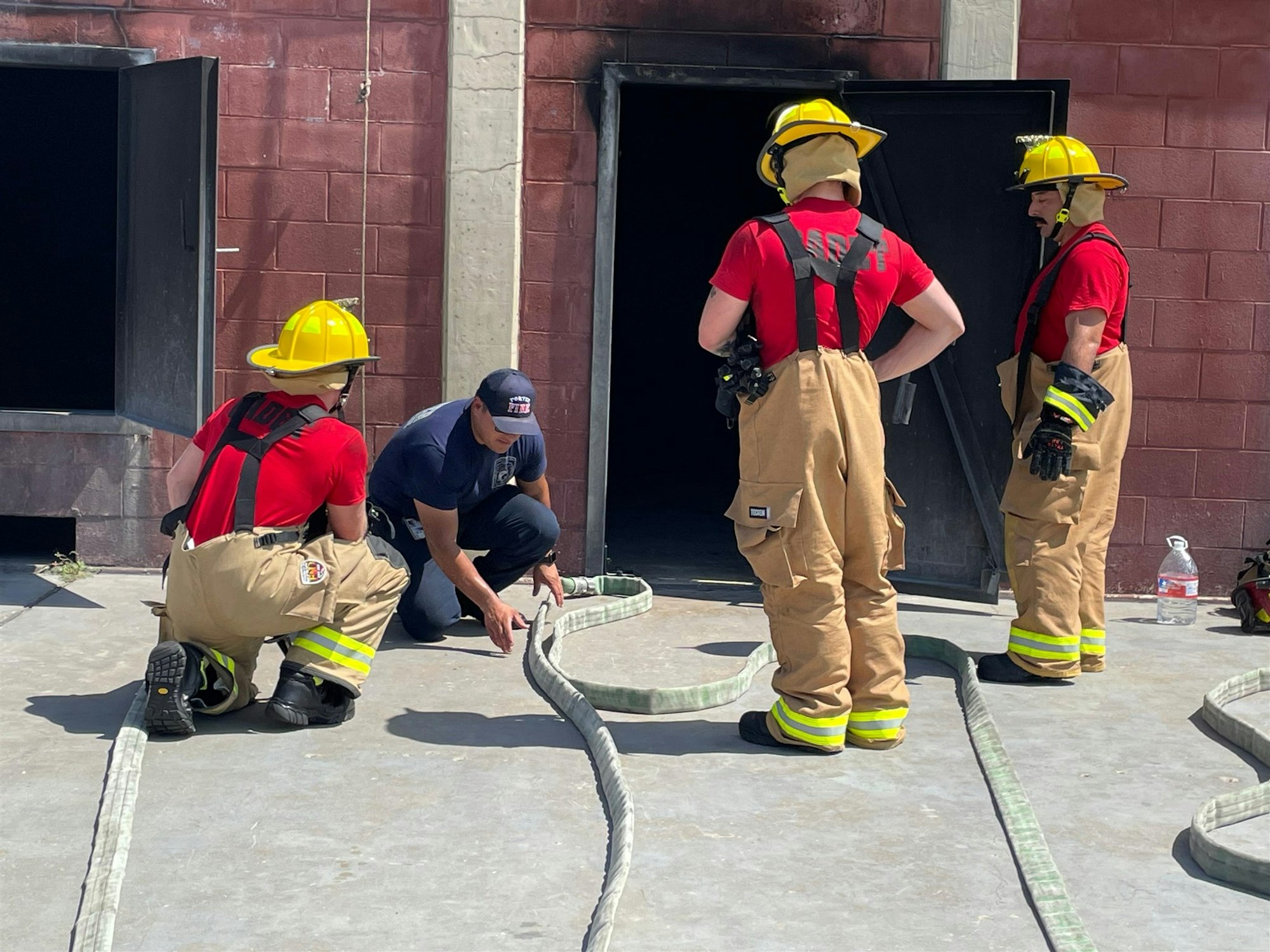 Firefighters in training are inspecting hoses outside a training facility, preparing for a drill or exercise.