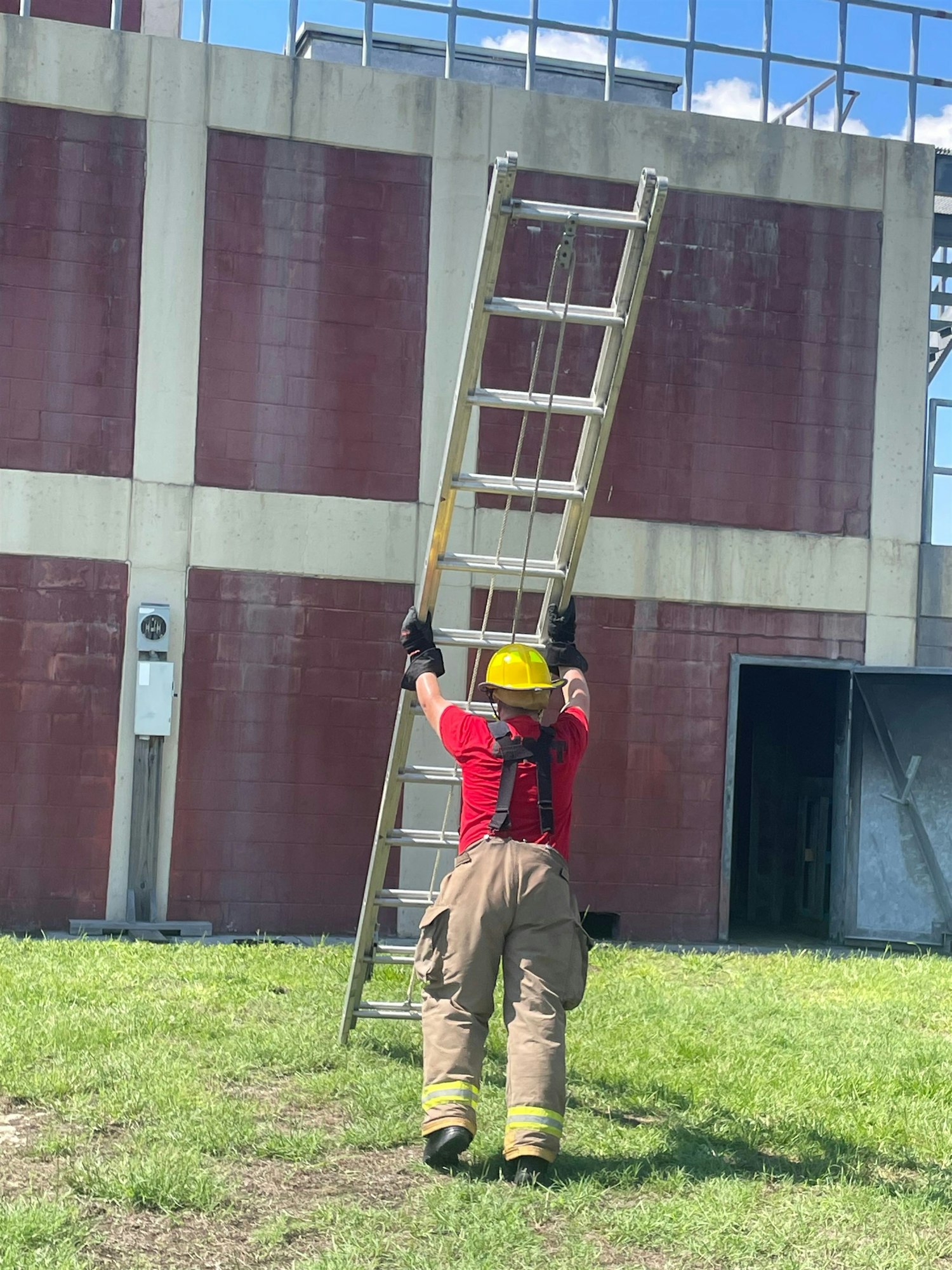 A firefighter in protective gear is lifting a ladder against a red wall, preparing for a training exercise or emergency response.