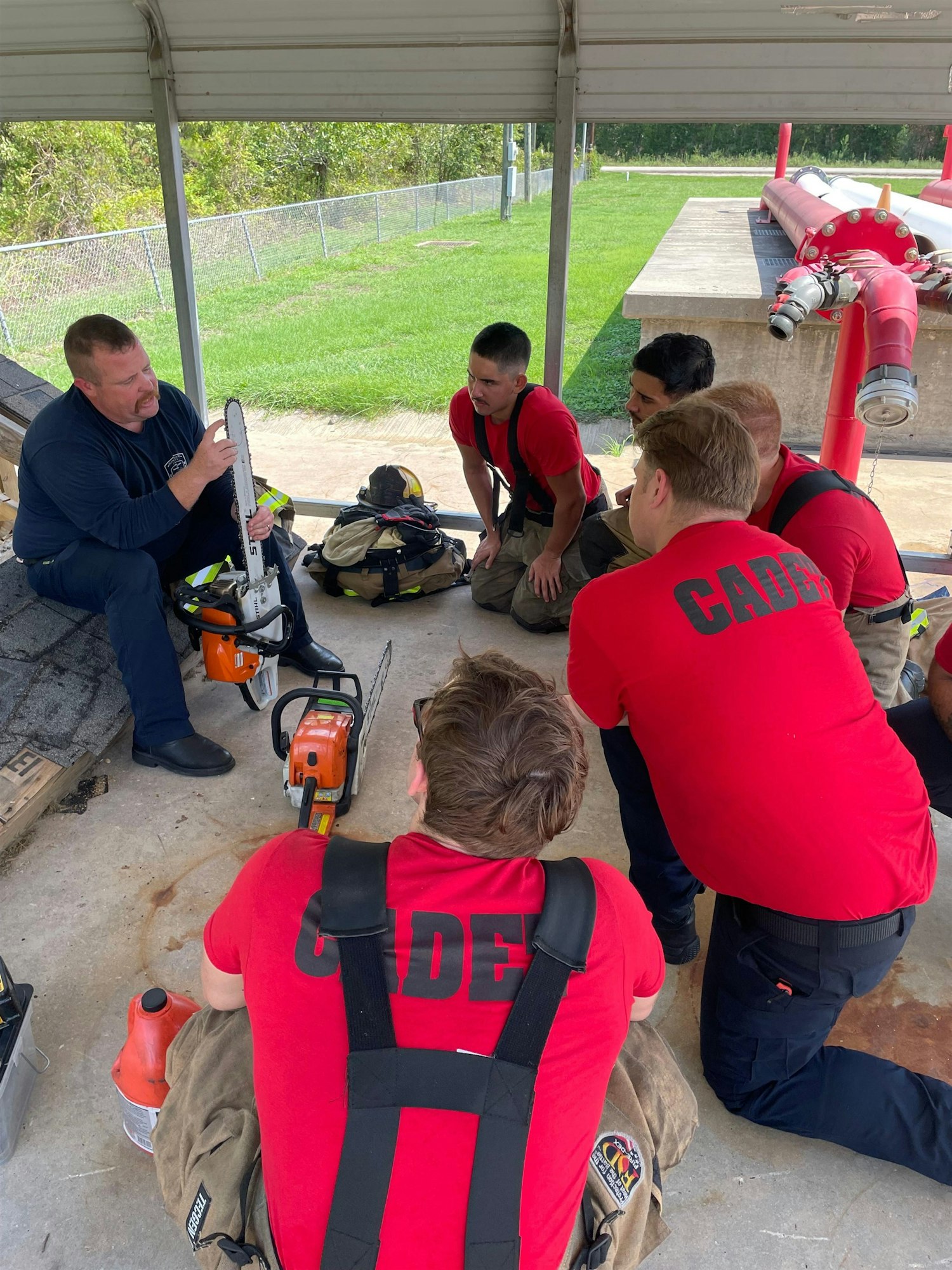 A group of fire cadets is listening to an instructor demonstrating how to use a chainsaw during training.