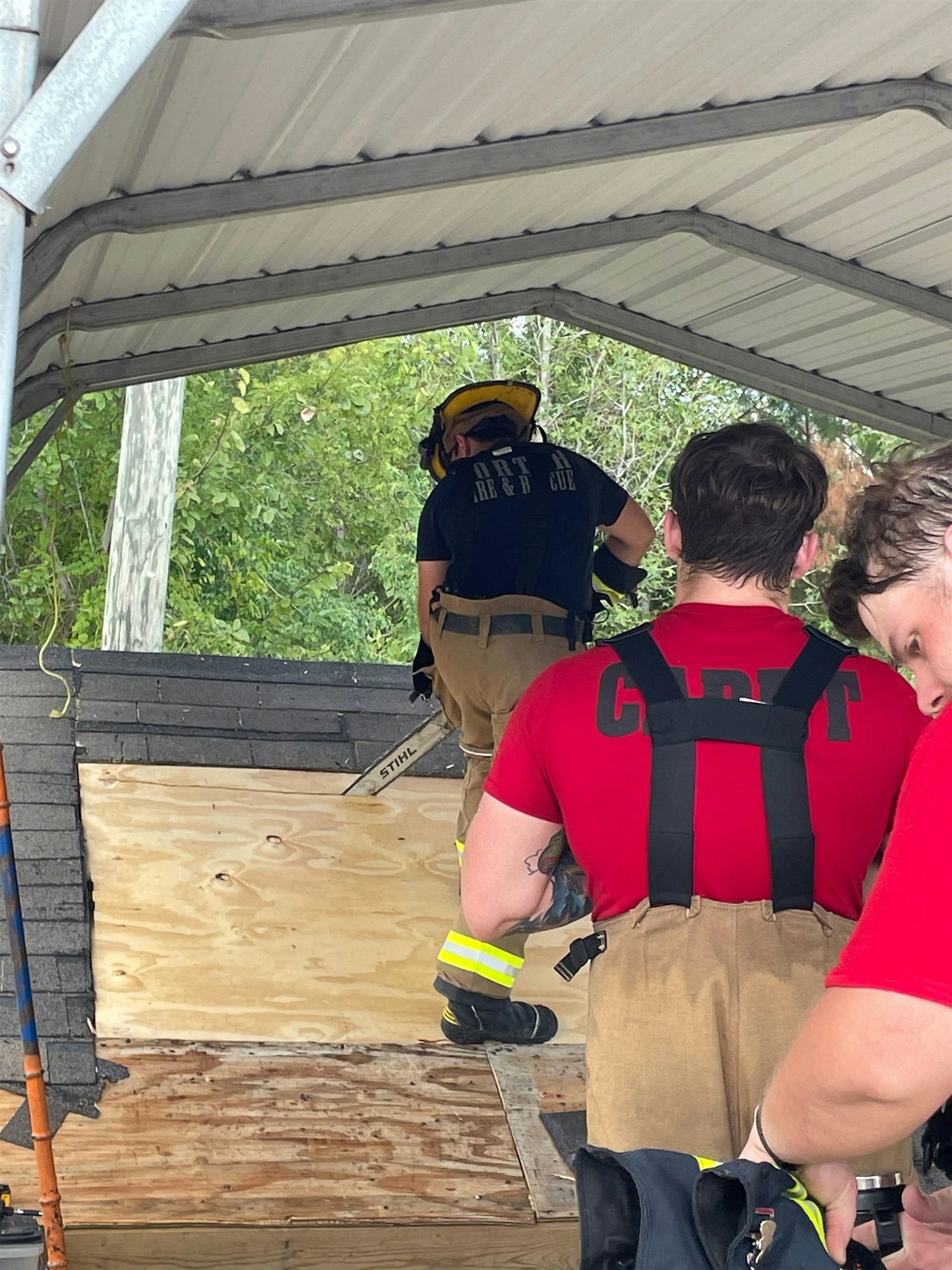Firefighters in training on a roof, using tools for practice, wearing gear and working on a structure under a canopy.