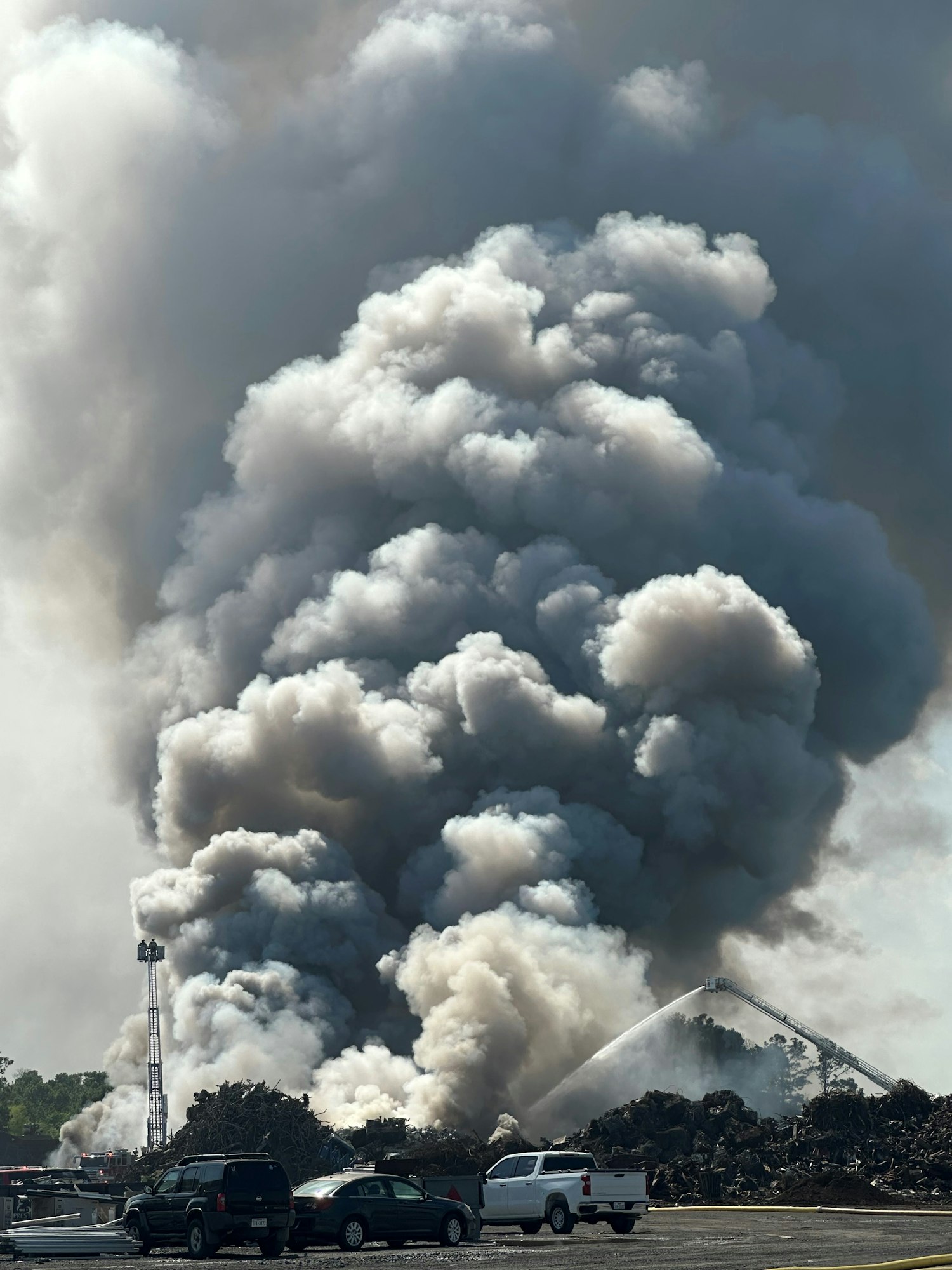 A large plume of smoke rises from a fire, as firefighters with a hose on an aerial ladder aim water at the blaze, vehicles in the foreground.