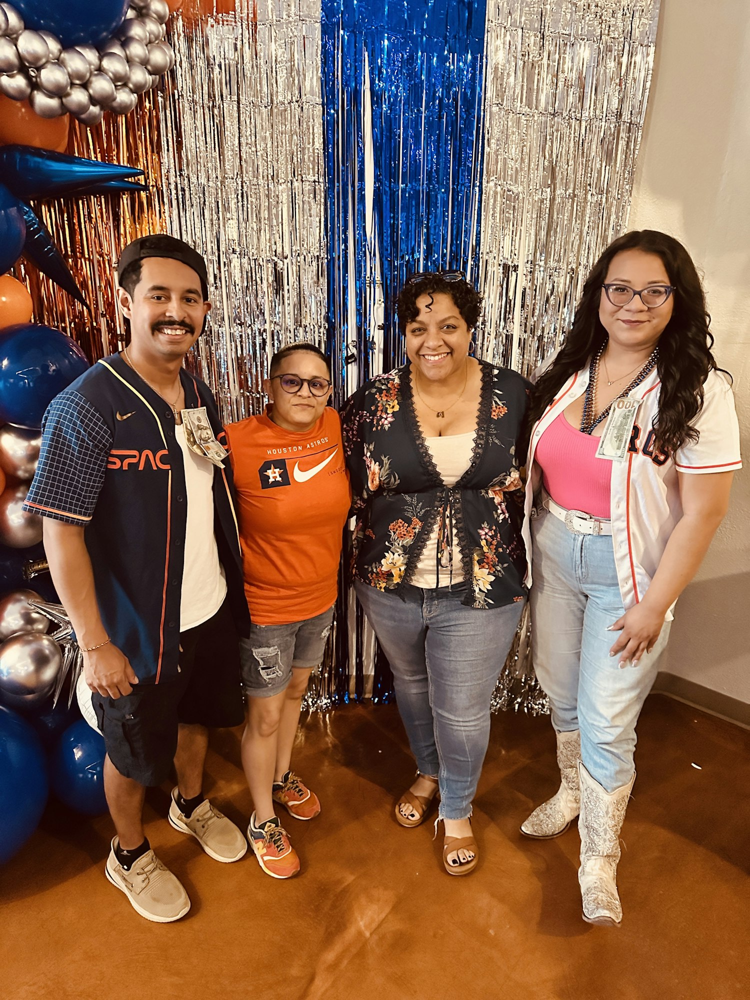 A group of four people poses together, smiling, in a festive setting with colorful decorations in the background.