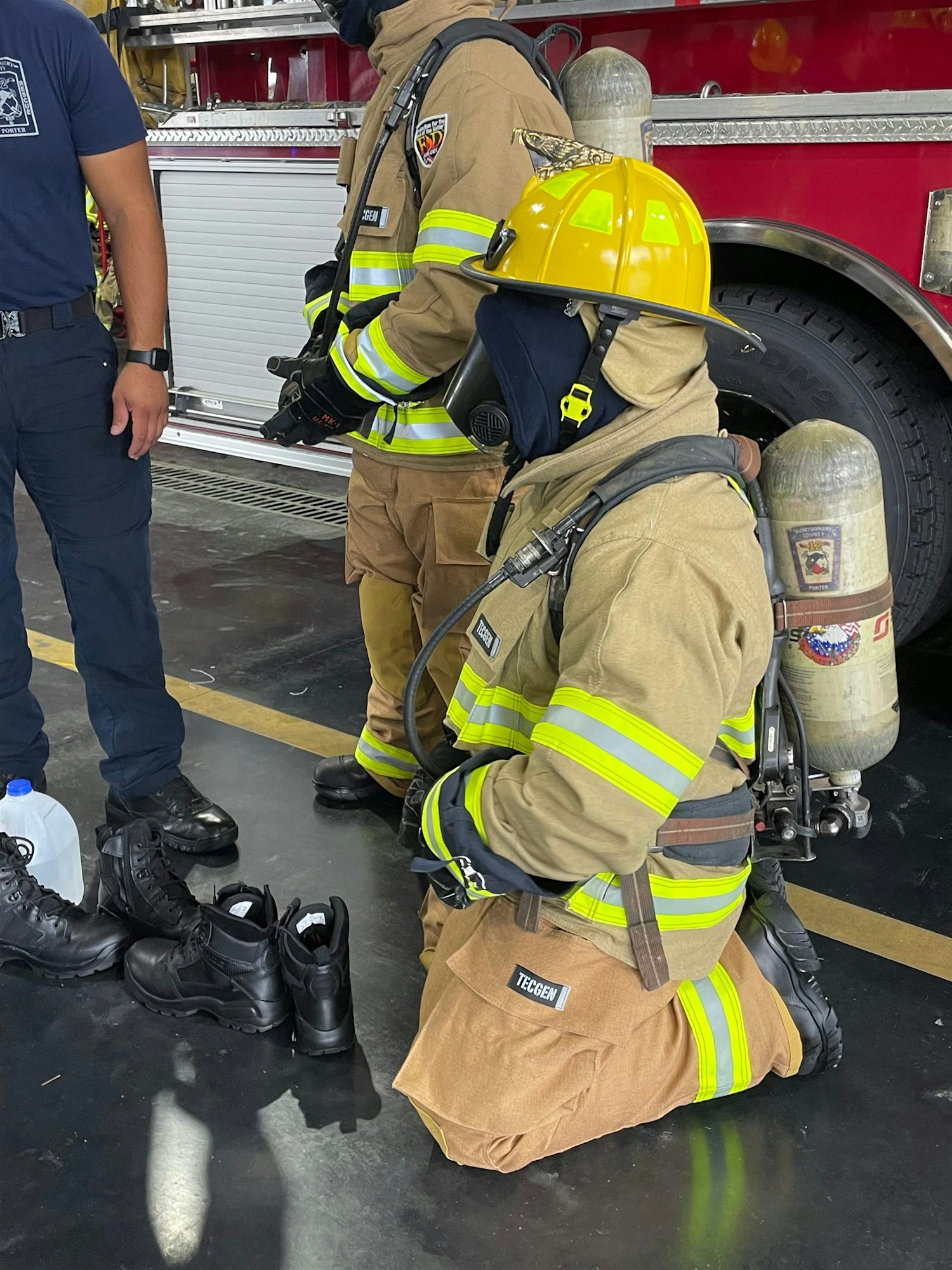 Two firefighters in gear are discussing in a fire station, with safety equipment and boots visible on the floor.
