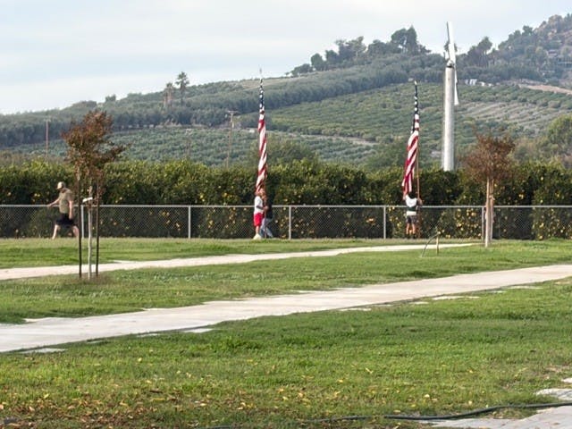 People walking by American flags, with a wind turbine and hilly landscape in the background.