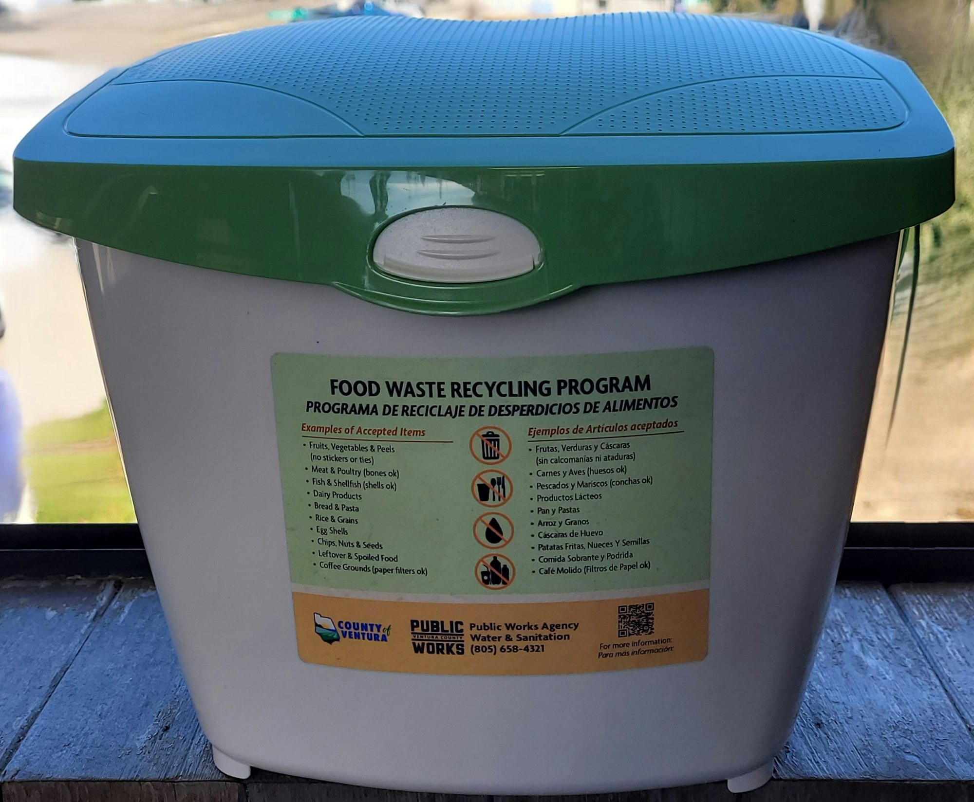 A green-lidded recycling bin with a list of accepted food waste items for a recycling program.