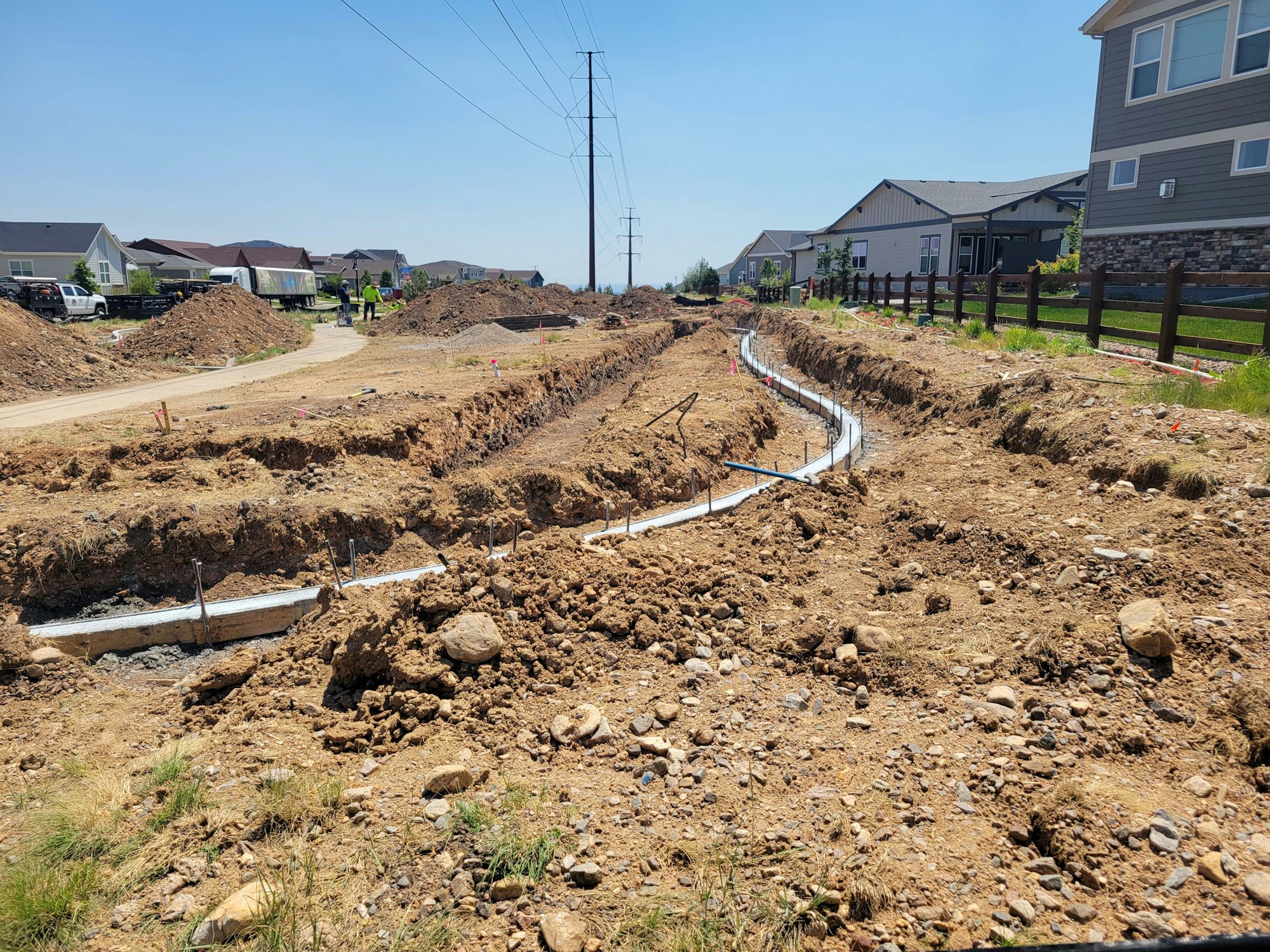 The image shows a construction site with trenches, dirt mounds, and utility lines, set against a backdrop of houses.