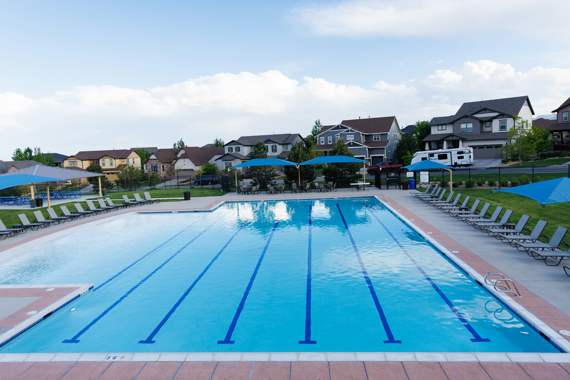 A community pool with clear water, lounge chairs, shaded areas, and houses in the background under a blue sky.