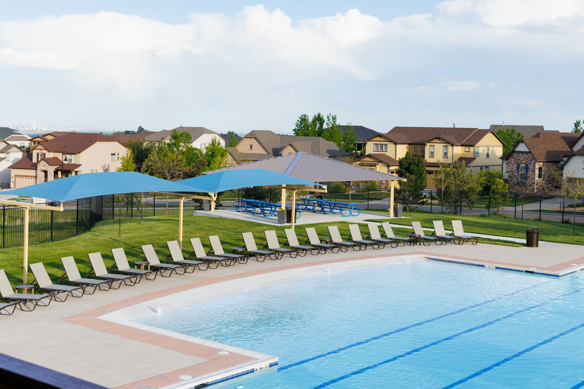 The image shows a swimming pool area with lounge chairs, shaded structures, and nearby residential houses. It's a sunny day.
