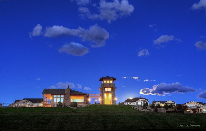 A twilight scene with a lit-up building featuring a tower, surrounded by houses under a sky dotted with clouds.