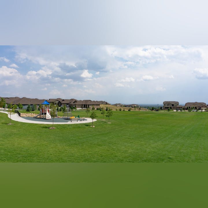 A spacious park with a playground and residential houses in the background, under a partly cloudy sky.