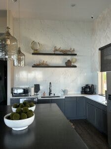 A modern kitchen with marble backsplash, floating shelves, and a bowl of limes on the counter.
