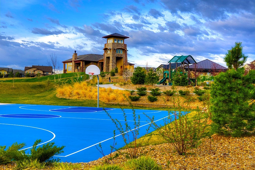 A colorful outdoor scene with a basketball court, playground, large house, and cloudy sky.