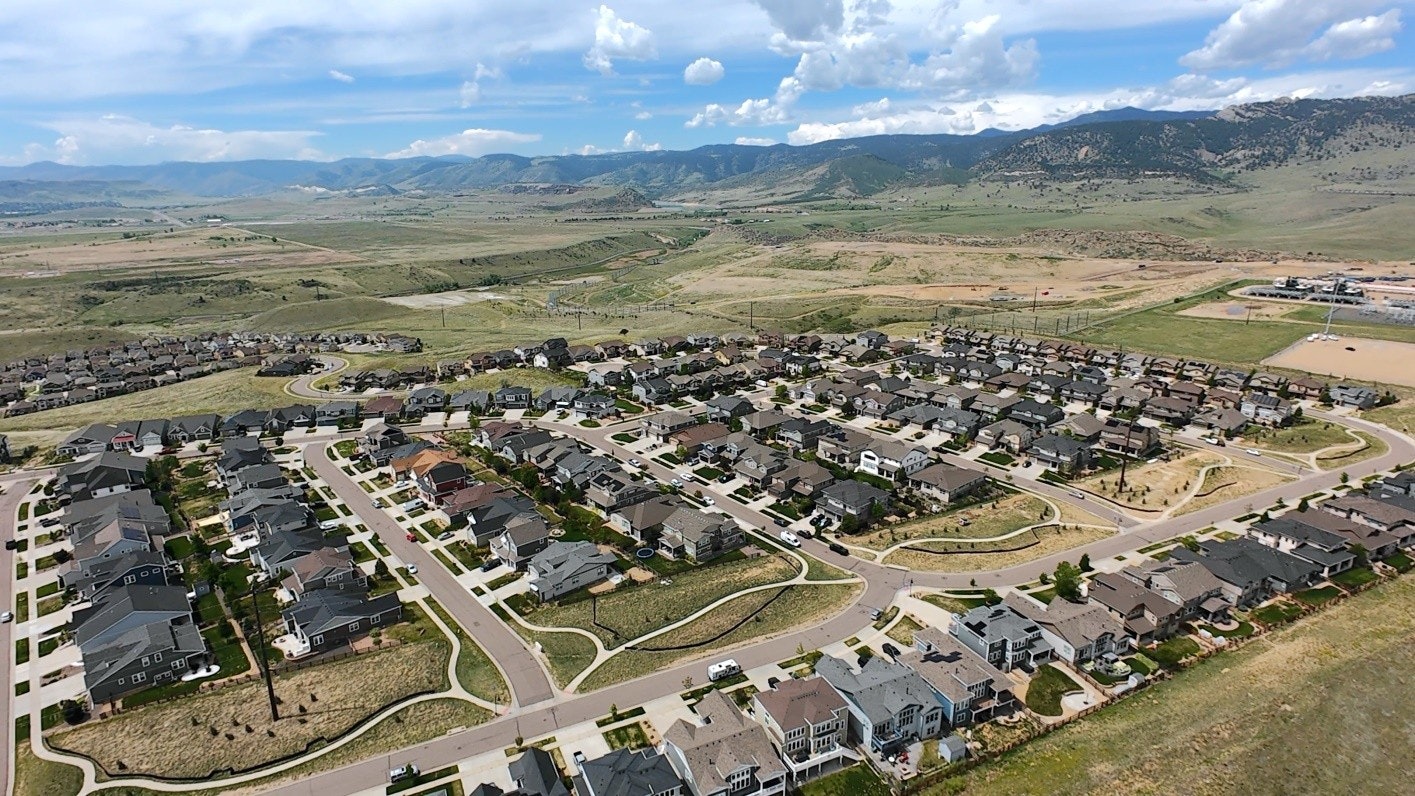 Aerial view of a suburban neighborhood with houses, winding streets, and mountains in the background. Green, open land nearby.