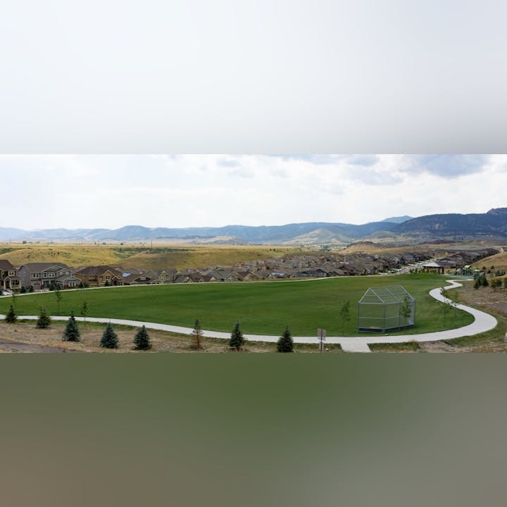 A suburban housing development with a large grassy area, walking paths, and a backdrop of rolling hills.