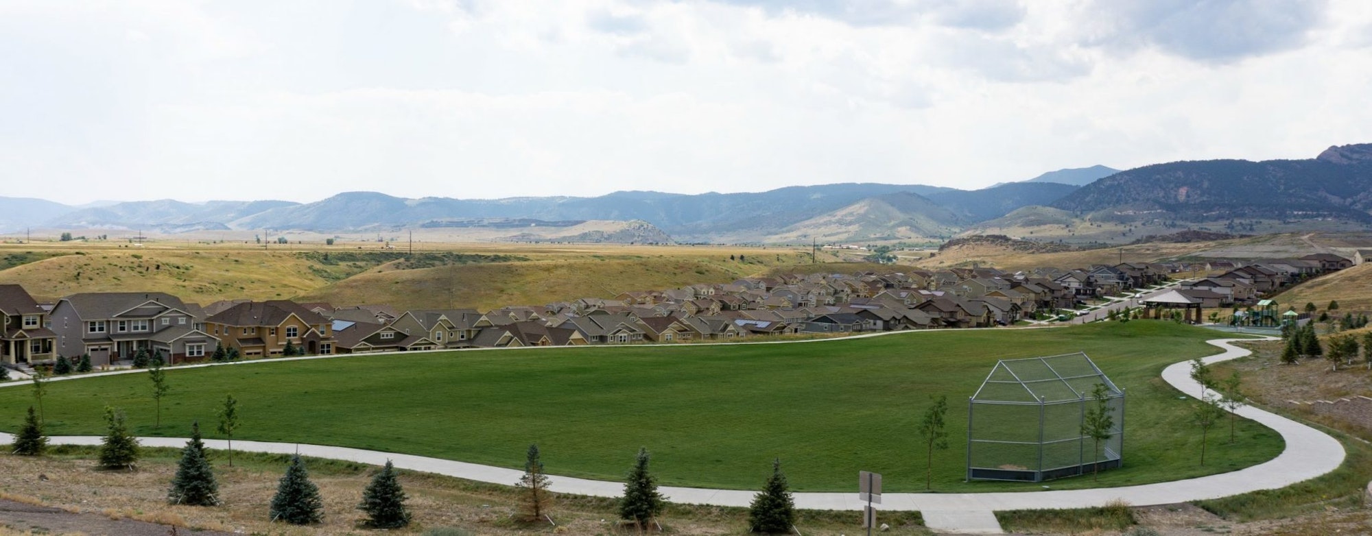 A suburban housing development with a large grassy area, walking paths, and a backdrop of rolling hills.
