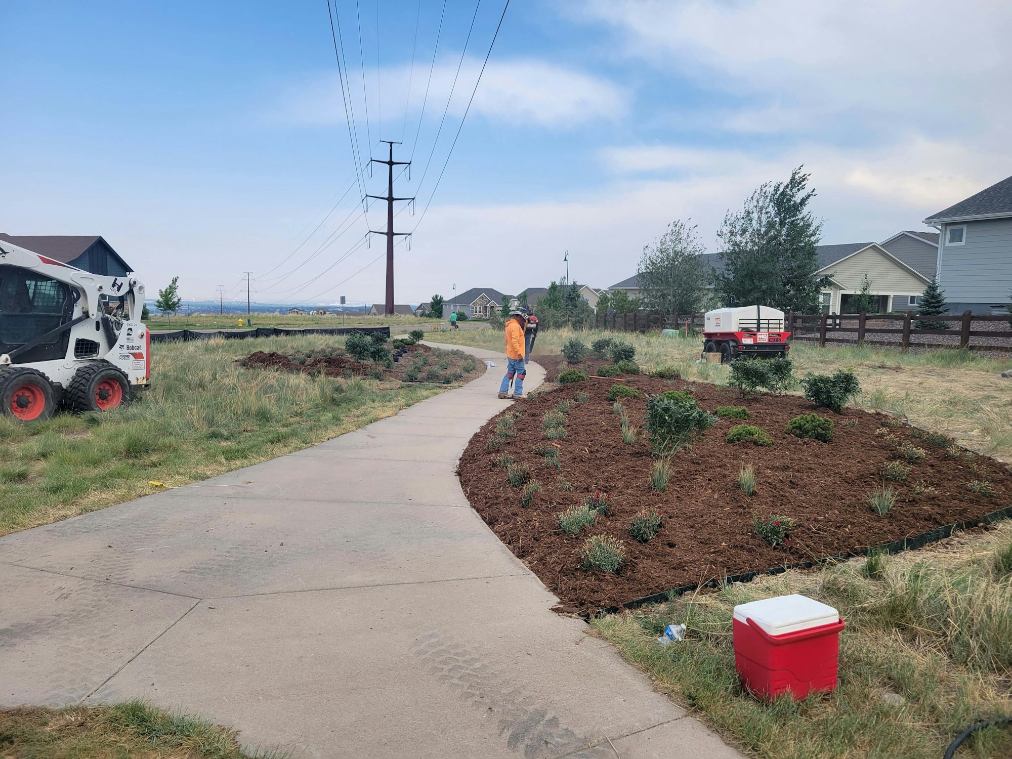 A pathway with landscaping work in progress, featuring a skid steer, workers, and newly planted shrubs and mulch.