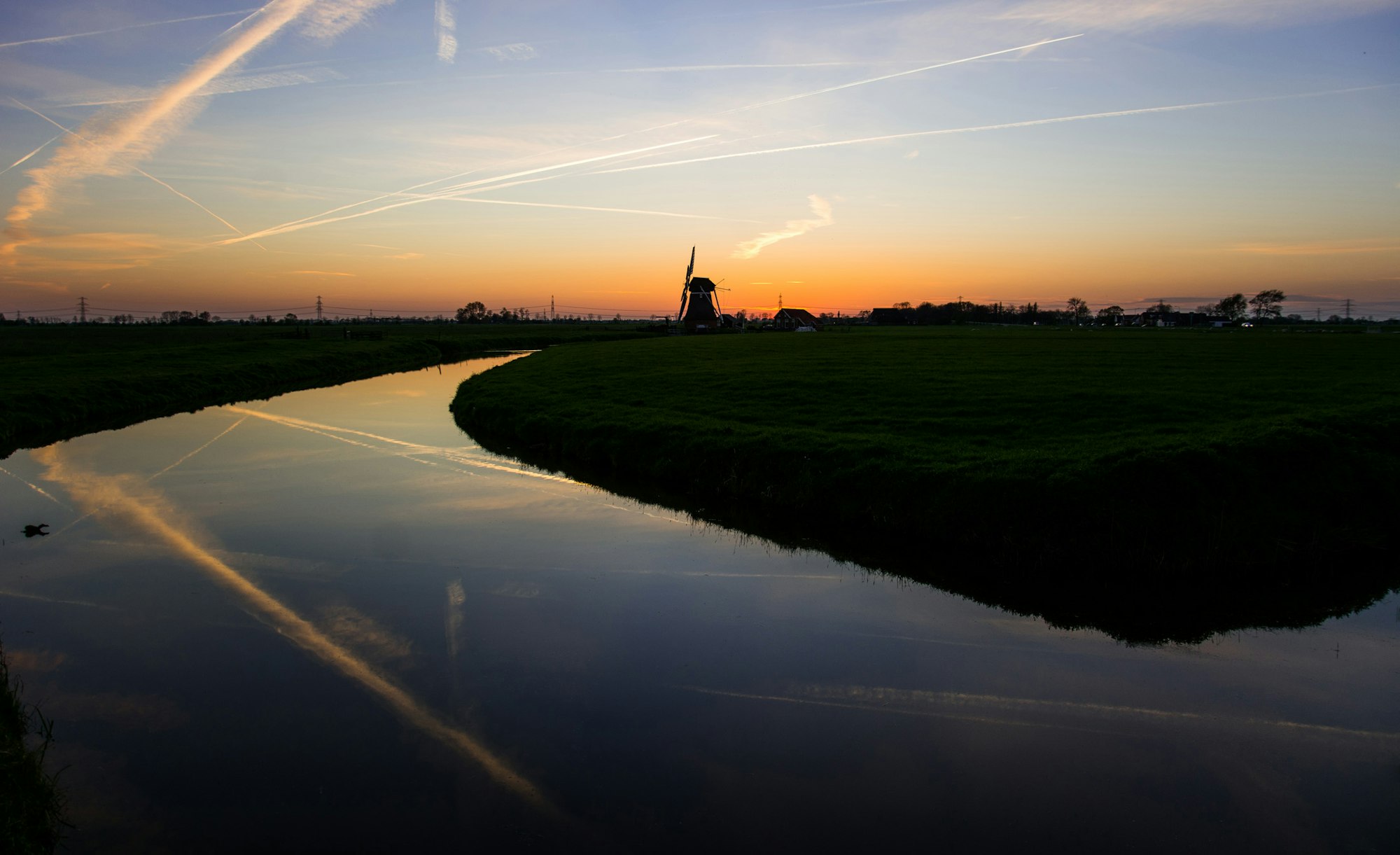 A tranquil sunset over a winding river reflecting the sky, with a windmill in the distance against lush green fields.