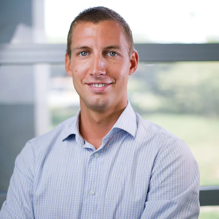 A smiling man in a checked shirt poses for a photo, with a blurred green background and large windows behind him.