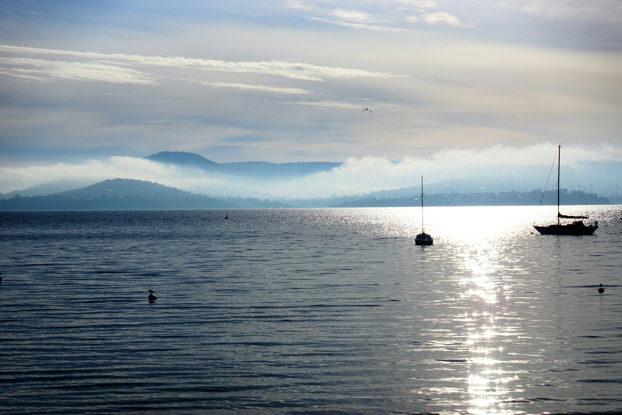 A serene view of a calm water body with sailboats, misty mountains in the background, and a cloudy sky reflecting sunlight.
