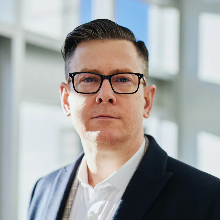 A person with glasses and a polished look, wearing a blazer, stands in a well-lit space with a neutral background.