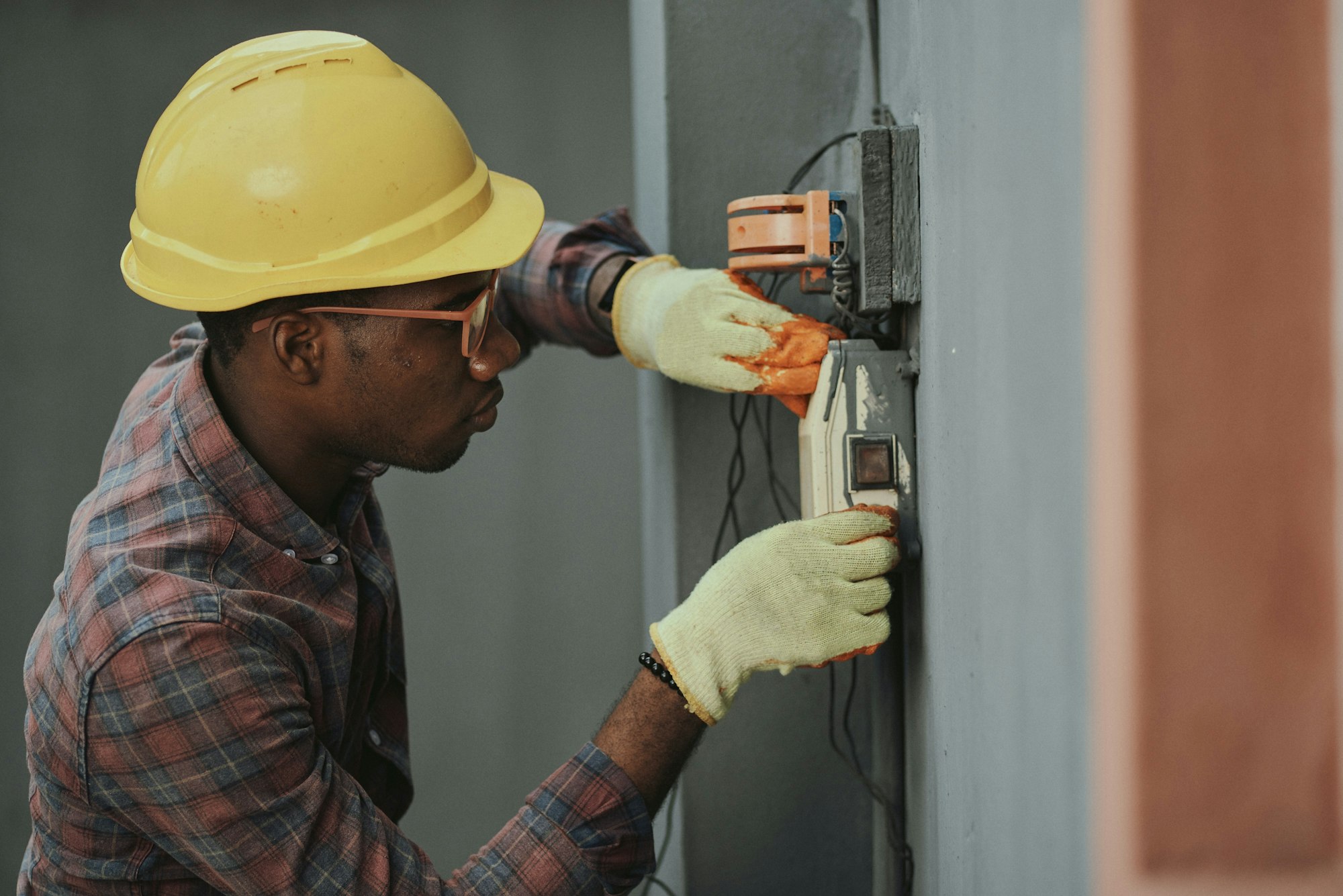 A worker in a yellow hard hat and gloves is inspecting and maintaining an electrical device on a wall.
