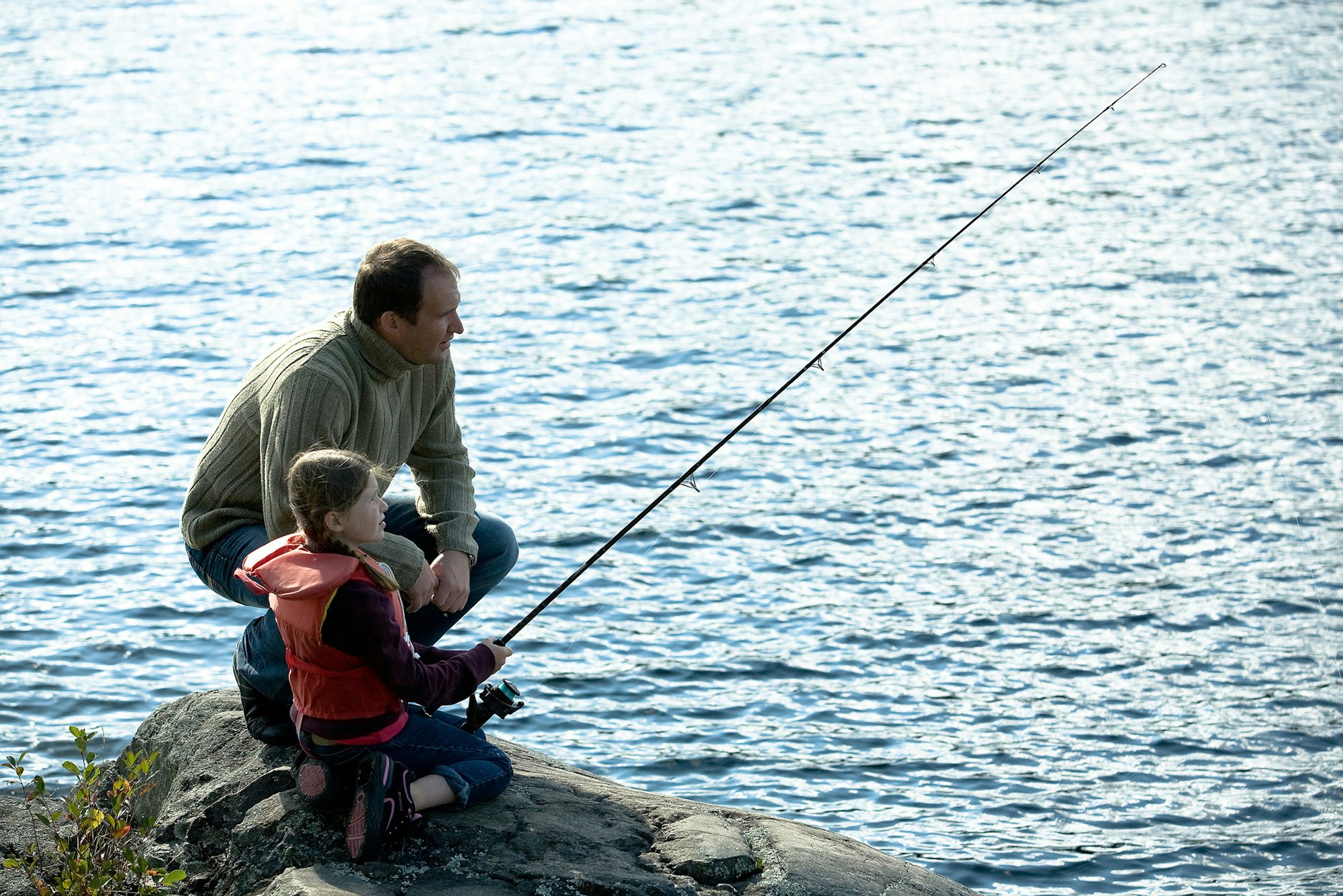 A man and a girl are fishing together by the water, enjoying a peaceful moment on a rock.