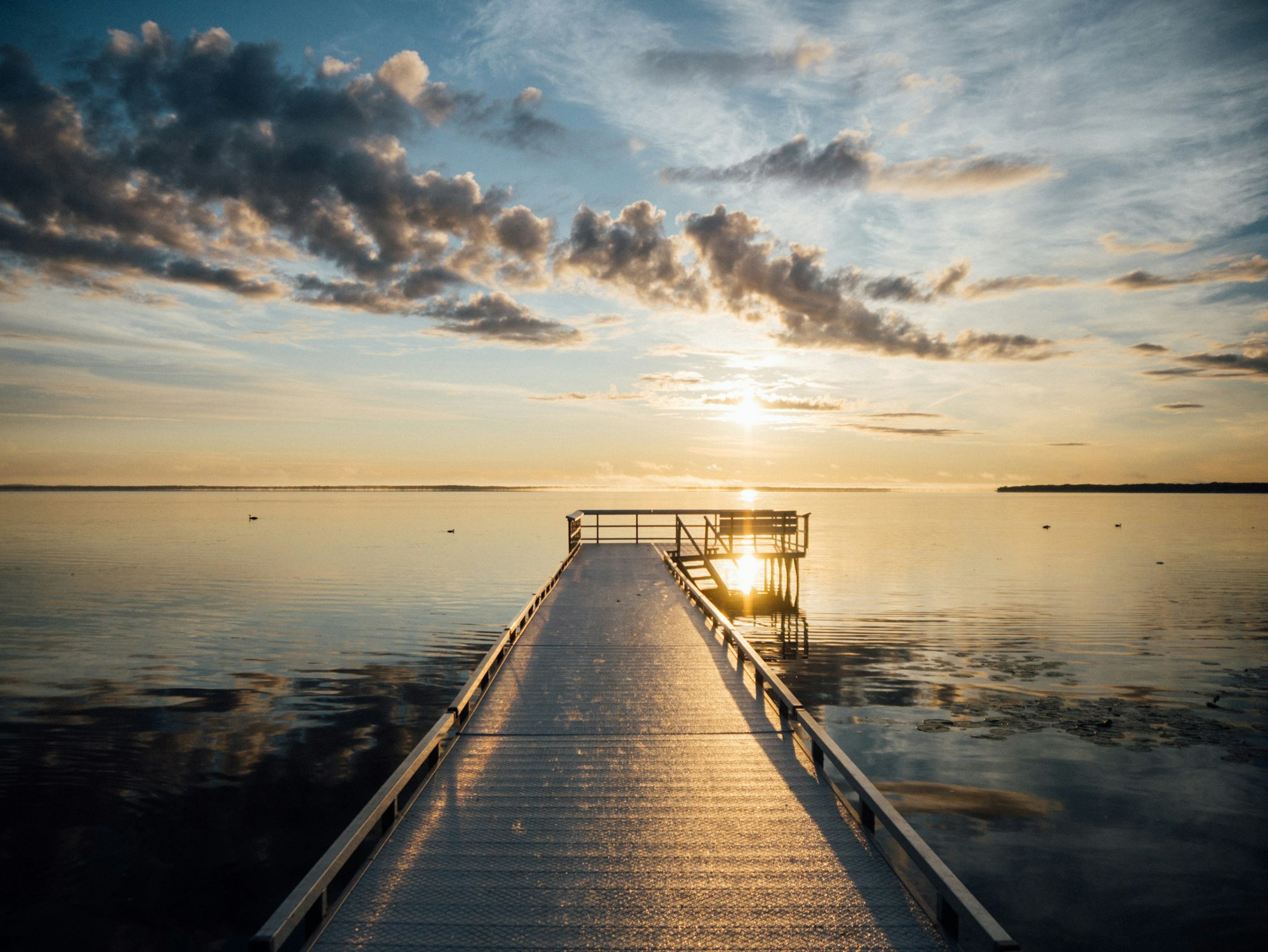 A serene dock extends into a calm body of water, reflecting golden sunlight and soft clouds during twilight.
