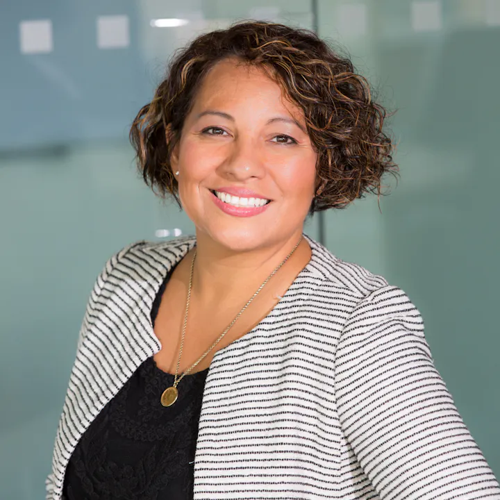 A smiling woman with curly hair, wearing a striped blazer and necklace, poses in an office setting with a glass background.